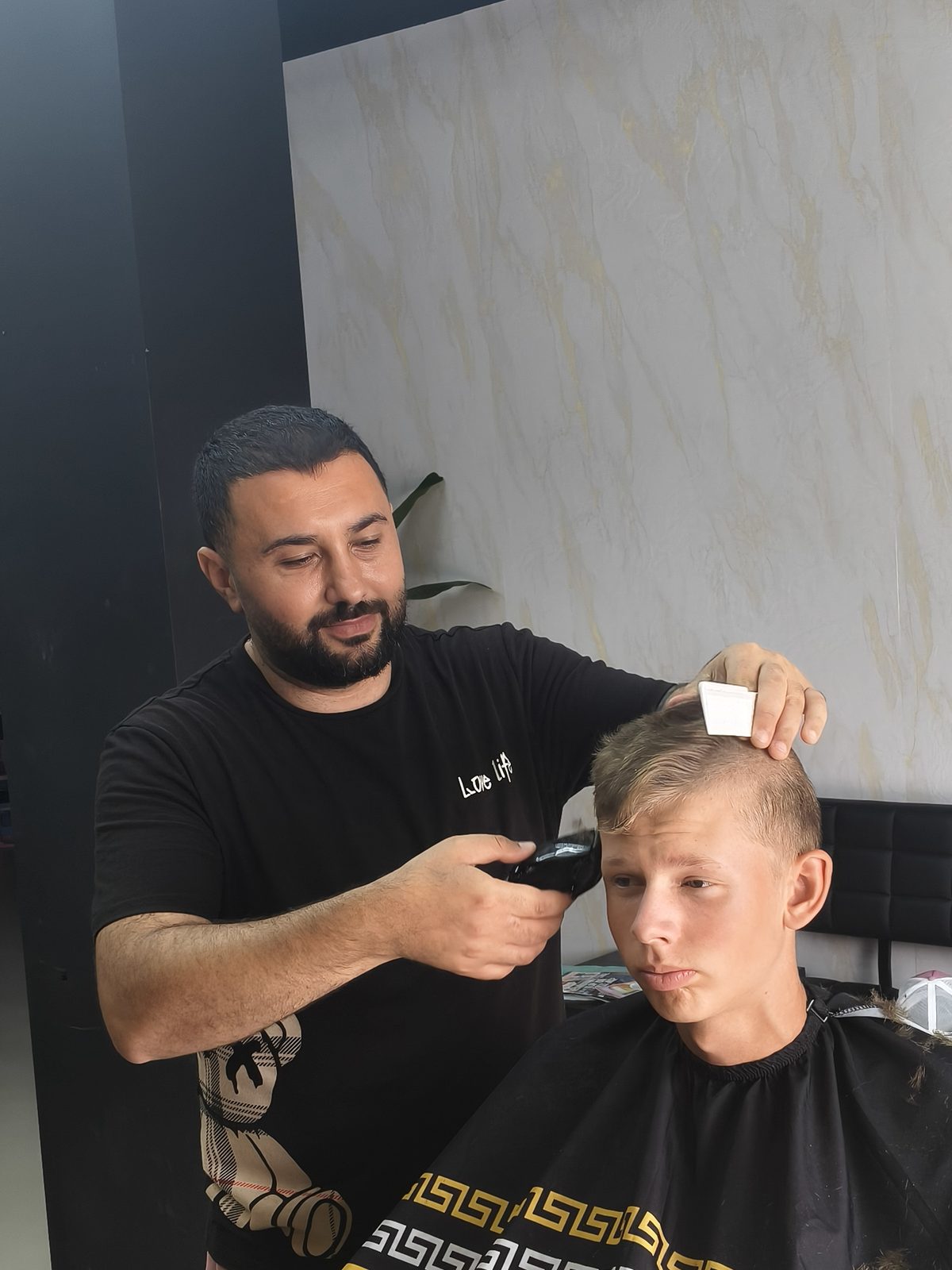 Een barber knipt met de tondeuse het haar van een jongen in de zwart met gouden cape